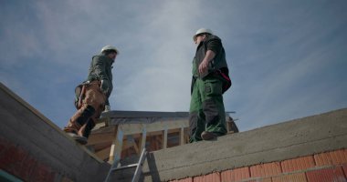 Çatıda projeyi tartışan iki inşaat işçisi, kask takıyorlar, konut inşaatı projesinde takım çalışması yapıyorlar, iletişim ve işbirliğine odaklanıyorlar.