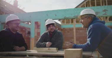 Üç inşaat işçisi inşaat alanındaki planları tartışıyor, başlık takıyor, çalışma masasında duruyor, sohbet ediyor, konut inşaatı projesiyle uğraşıyor.