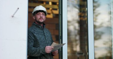 İnşaat işçisi elinde planlar, odaklanarak yukarı bakıyor, başlık takıyor, tamamlanmamış binanın kapısında duruyor, planlama ve mimari teftişi temsil ediyor.