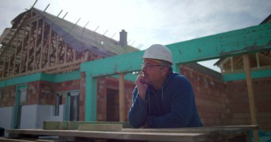 İnşaat işçisi inşaat alanındaki projeyi, düşünceli erkek inşaat işçisini, arka planda inşaat halindeki konut yapısını yansıtıyor.