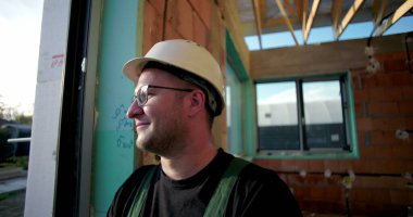 İnşaat işçisi kasketli, gülümseyen, dışarıdan bakan, yan profilli, doğal çevresi olan, inşaat alanında rahat tavırlı biri.