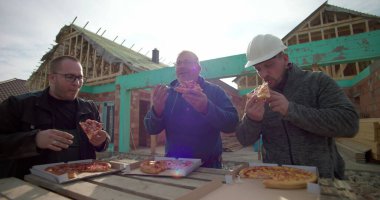 İnşaat işçileri inşaat alanında pizzanın tadını çıkarıyorlar, dinlenmek için biraz zaman ayırıyorlar, arka planda tamamlanmamış bina yapısı, açık havada gündelik yemek.