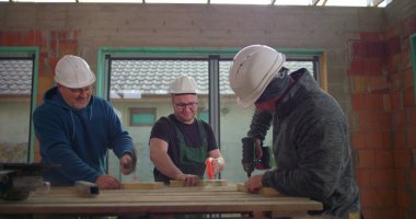 Ahşap işlerinde işbirliği yapan üç inşaat işçisi, ahşap masa üzerinde matkap ve aletler, inşaat alanında takım çalışması, güvenlik kaskları, kapalı alanda yenileme