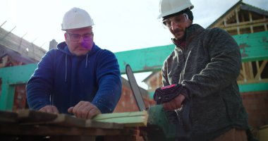 İnşaat alanında elektrikli testereyle çalışan iki inşaat işçisi, inşaat halindeki konut yapısı, hassas ahşap kesimi, çerçeve çalışmasında takım çalışması