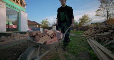 Kısmen inşa edilmiş evin önüne tuğla dolu el arabasını iten inşaat işçisi, dış duvara da yalıtım malzemesi yerleştiriyor.
