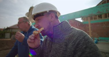 İnşaat işçilerinin iş yerinde öğle yemeği arasında pizzanın keyfini çıkardıkları yakın plan çekimler, dostluk ve rahatlama, arka planda tamamlanmamış bina yapısı, gündelik yemek