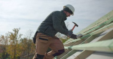 İnşaat işçisi inşaat yapısına çatı kirişleri çakıyor, güvenlik kaskı takıyor, arka planda iş arkadaşlarıyla takım çalışmasının yüksek açılı görüntüsü, açık hava inşaat projesi sitesi