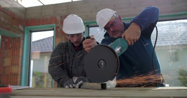 Metal üzerinde elektrikli öğütücü kullanan inşaat işçileri kıvılcımlar üretiyor, iş başında takım çalışması yapıyor, güvenlik kaskı ve koruma için gözlük takıyor, yenileme sahasını yeniliyor.