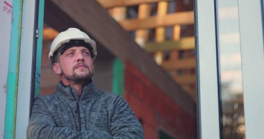 Kapıda inşaat işçisi, kollar çapraz, kasketli yana bakıyor, bina arka planı tamamlanmamış, kendini adamışlık, profesyonellik ve özen gösterme