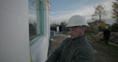 İnşaat işçisi inşaat alanında mezura ile duvarı ölçüyor, doğruluğa odaklanıyor, başlık takıyor, yeni yapı için temel kuruyor, açık hava düzeneği kuruyor.