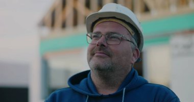 Sert şapkalı inşaat işçisi düşünceli bir ifadeyle bir inşaat projesinde, açık havada bir yere bakıyor.