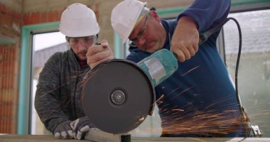 Açı öğütücüyle metali kesen iki inşaat işçisi, yoğun kıvılcımlar, odaklanmış takım çalışması, güvenlik kaskları ve yenileme alanındaki gözlükler.