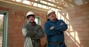 İki inşaat işçisi, kısmen inşa edilmiş bir binada güven içinde duruyor, kasketli şapkalar takıyor ve gülümsüyor, şantiyede takım çalışması ve güven gösterisi yapıyorlar.