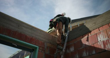 Çatıdan merdiven inen inşaat işçileri, kullanımda olan güvenlik ekipmanları, inşaat alanında dikkatli adımlar, endüstriyel takım çalışması, mavi gökyüzünün altındaki bina