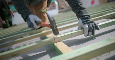 İnşaat işçilerinin çatı kirişlerine çivi çakması, güvenlik eldiveni takması, el ve çekice odaklanması, arka planda meslektaşları olan açık hava inşaat projesi.