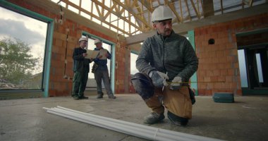 İnşaat işçisi, binanın iç kısımlarındaki PVC borularını diz çöküp ölçerken, arka plandaki meslektaşları da projeleri, inşaat alanındaki takım çalışmalarını gözden geçiriyorlar.