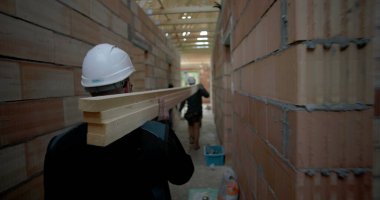 İnşaat işçisi omuzunda tahta kalaslar taşıyor, bitmemiş bir binada dar koridorlarda yürüyor, inşaat alanında takım çalışması ve işçilik yapıyor.