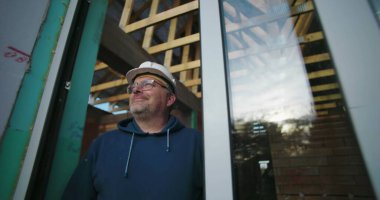 Daha yaşlı bir inşaat işçisi. Camdan dikkatlice bakıyor. Ahşap kirişlerle çerçevelenmiş.