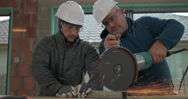 Metali elektrikli öğütücüyle kesen iki inşaat işçisi, yoğun kıvılcımlar uçuşuyor, güvenlik teçhizatı açık, takım çalışması ve yenileme projesine odaklanıyor