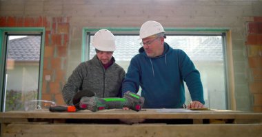 İki inşaat işçisi ahşap bir masanın üzerine bina planlarını, yenileme projesinde takım çalışmasını, ön planda aletleri, kapalı alanda takılan güvenlik kasklarını tartışıyor.