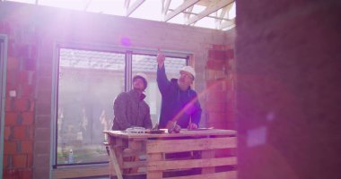 İnşaat işçileri inşaat alanındaki ahşap masa üzerinde öğütücü alet kullanıyorlar, takım çalışması ve işbirliği, güvenlik kaskları, yenileme projesi üzerine odaklanmış tartışmalar,