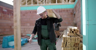 İnşaat işçisi bir yığın ahşap kalas taşıyor, inşaat sahasında çerçeve yapmak için malzeme hazırlıyor, konut inşaatına odaklanıyor, marangozlukta takım çalışması yapıyor.