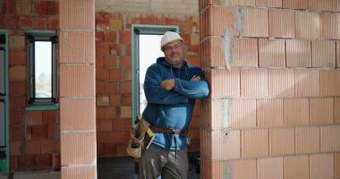 Mavi kapüşonlu gülümseyen inşaat işçisi, kolları bağlı, tamamlanmamış binadaki tuğladan duvara yaslanmış, güvenlik kaskı takılı, sanayi bölgesi arka planı