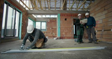 Bitmemiş binanın zeminindeki PVC borularını ölçen inşaat işçisi, iki meslektaşı arka plandaki planları, takım çalışmasını ve inşaat alanındaki odak noktalarını tartışıyor