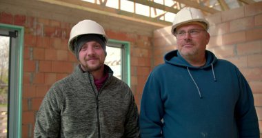 İki inşaat işçisi, kısmen inşa edilmiş bir binada güven içinde duruyor, kasketli şapkalar takıyor ve gülümsüyor, şantiyede takım çalışması ve güven gösterisi yapıyorlar.