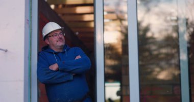 İnşaat işçisi kapı eşiğine dayanmış, kollar çapraz, başlık takmış, kendinden emin ve düşünceli görünüyor, bina projesini denetliyor, profesyonel duruş.