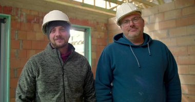 İnşaat halindeki bir binanın içinde gülümseyen iki inşaat işçisi. Birinin kolları bağlı, kısmen inşa edilmiş bir yapıda takım çalışması ve kendine güveni var.