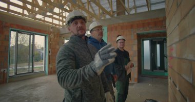 İnşaat işçisi duvarı işaret ederken, inşaat ortamında proje ayrıntılarını, takım çalışmasını ve iletişimi de anlatıyor