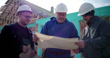 İnşaat alanındaki inşaat planlarını inceleyen üç inşaat işçisinden oluşan bir grup, proje detaylarını arka planda tamamlanmamış bir yapıyla tartışıyor.