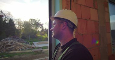 Dışarıya bakan inşaat işçisi, gülümseyen, yandan bakan, inşaat enkazı ve arka planda ağaçlar olan, gevşek ifadeli.