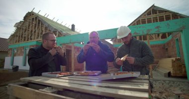 İnşaat işçileri inşaat alanında pizzanın tadını çıkarıyorlar, dinlenmek için biraz zaman ayırıyorlar, arka planda tamamlanmamış bina yapısı, açık havada gündelik yemek.