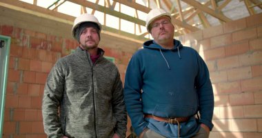 İnşaat alanında kapalı alanda poz veren iki inşaatçı, güvenlik kasklı adamlar takım çalışması ve güven gösteriyor, tamamlanmamış tuğla yapıda çalışıyorlar, ilerleme kaydediyorlar.