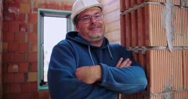 İnşaat işçisi inşaat halindeki tuğladan duvara yaslanıyor, kollar çapraz, güvenlik kaskı takıyor, rahat ve kendinden emin duruş, endüstriyel ortam.