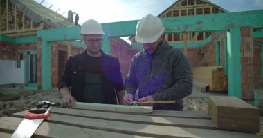 Bir inşaat sahasında ahşabı ölçüp işaretleyen iki müteahhit, takım çalışması ve hassasiyet, konut projesi, yeni bir ev, sağlam şapkalar, işe yarar araçlar.