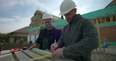 Tecrübeli müteahhitler bir inşaat sahasında ahşabı kalem ve mezurayla ölçüp işaretliyor, takım çalışması, hassasiyete, güvenlik kasklarına odaklanıyor, arka plandaki inşaat binasının altında.