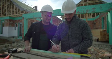 Ahşabı hizalayan ve ölçen iki müteahhit, inşaat alanında takım çalışması, yeni ev iskeleti projesi, konut yapısında işbirliği, sert şapkalar takmak.