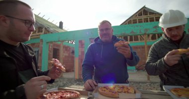 İnşaat işçileri inşaat alanında pizzanın tadını çıkarıyorlar, dinlenmek için biraz zaman ayırıyorlar, arka planda tamamlanmamış bina yapısı, açık havada gündelik yemek.
