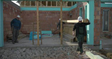 İnşaat alanında omuzlarında ahşap kalaslar taşıyan işçiler, takım çalışması ve marangozluk görevlerine odaklanmış durumda, görünür yapı ve kirişlerle yerleşim alanı inşaatı sürüyor