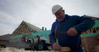 Ahşap bloğa çivi çakan inşaatçı, göreve odaklanmış, derme çatma tezgahın yanındaki testere, arka planda tamamlanmamış bina yapısı olan inşaat alanı.