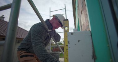 Dış duvardaki yalıtım panelini ruh seviyesine ayarlıyorum. İnşaat işçisi ev işleriyle uğraşıyor. Tuğla ve yalıtım düzenini kapatıyor.