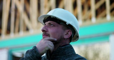 Elleri çenesinde inşaat işçisi, inşaat projesini düşünüyor, arka planda ahşap çerçeve yapısı, planlama ve problem çözmeyi sembolize ediyor.