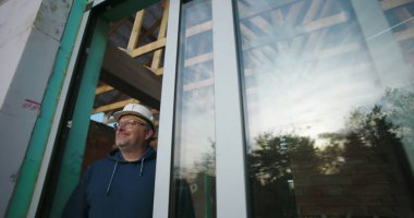 Daha yaşlı bir inşaat işçisi. Camdan dikkatlice bakıyor. Ahşap kirişlerle çerçevelenmiş.