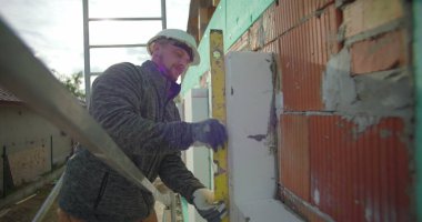 İzolasyon paneli ruh seviyesiyle karşılaştırılıyor. İnşaat işçisi yapı iskelesinde güvenlik ekipmanlarıyla çalışıyor. Duvarın dış duvarına tam oturmasını sağlıyor.