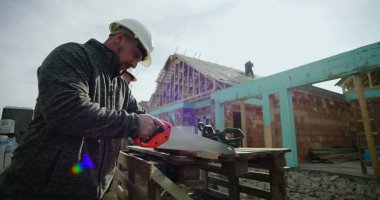 Ahşap kesmek için el testeresi kullanan, odaklanmış ve titiz, başlık takan, arka planda ev iskeleti olan inşaat alanı, yapı iskeleti üzerinde çalışan bir inşaat işçisi.