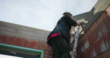 İki inşaat işçisi inşaat alanında merdiven çıkıyor, çatı yapısını güvence altına alıyor, güvenlik ve kalifiye işçilere odaklanıyor, konut inşaatı projesi devam ediyor