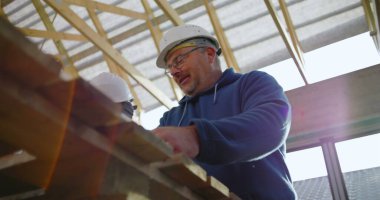 Tahta tutan inşaat işçisinin düşük açılı görüntüsü, yapısal projede takım çalışması, koruyucu kask, odaklı ifade, görünür yapı iskeleti.